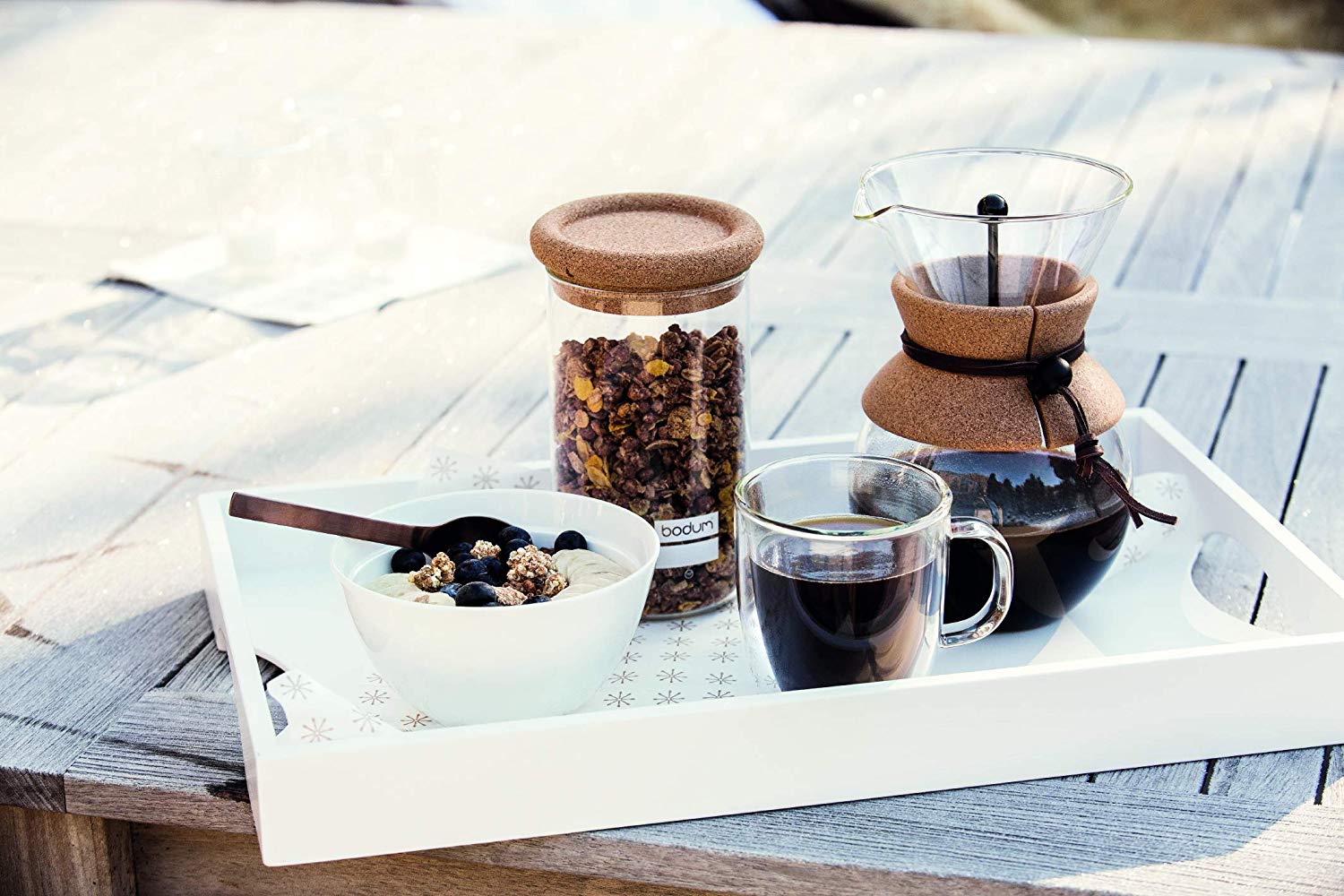 A tray with a coffee mug filled with coffee, coffee press, coffee grounds in a glass jar with a cork lid and a bowl of oatmeal.
